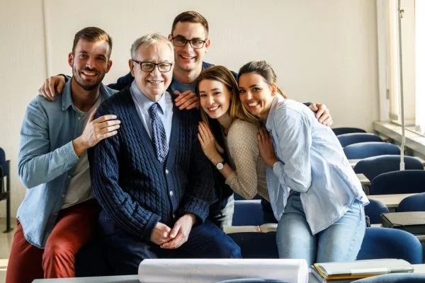 Smiling multi-generational group in a classroom, with an older man seated at the center surrounded by four younger adults showing support and connection—symbolizing mentorship, legacy, and family business succession.